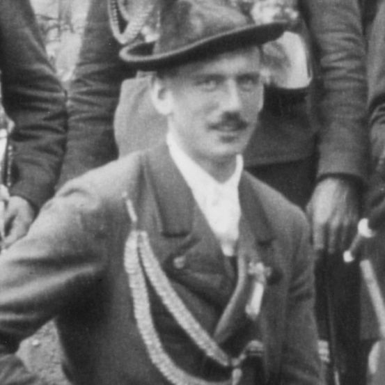 Schwarz-Weiß-Foto eines Mannes in Uniform, der einen Hut und eine Schärpe trägt, steht mit anderen Personen in einer formellen Umgebung.