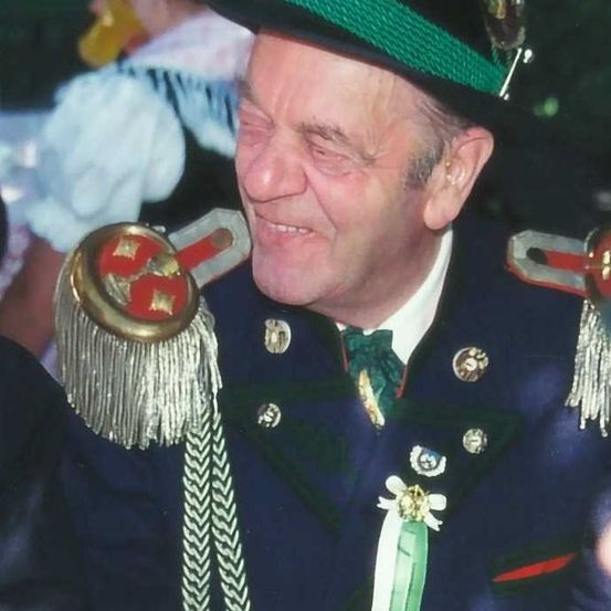 Ein älterer Mann in traditioneller bayerischer Tracht lächelt, trägt einen Hut mit grünem Band, ein golden verziert Accessoire und eine grüne Krawatte mit Silberverzierungen.