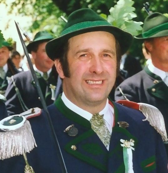 Ein lächelnder Mann in einem grünen Hut mit einem Schwert und dekorativen Federn. Hinter ihm stehen Menschen in grünen Hüten und Uniformen.