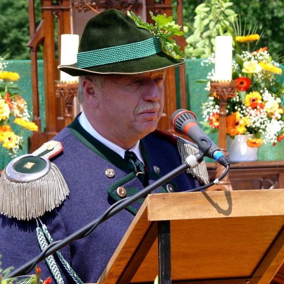 Ein Mann in traditioneller Uniform steht an einem Rednerpult und hält eine Rede. Er trägt einen grünen Hut mit grüner Einfassung und Blättern. Ein Mikrofon steht vor ihm. Dahinter befindet sich ein hölzerner Altar mit einer Kerze und Blumen.