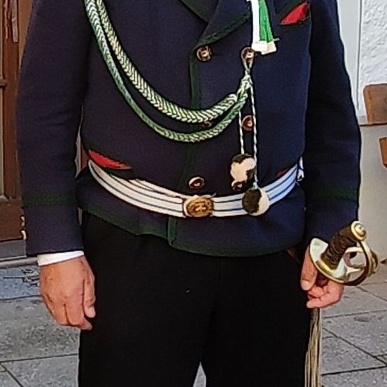 Ein Mann in traditioneller Militäruniform mit dekorativen Schulterschnüren und einem Schwert. Die Uniform ist blau mit grünem und rotem Besatz.
