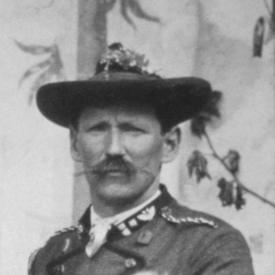 Ein altes Foto eines Mannes in Militäruniform mit einem Hut, der mit einer Blume verziert ist, vor einem verschwommenen Hintergrund.