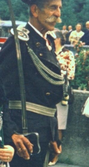 Ein älterer Mann in einer Militäruniform mit einem Schwert, einem Hut und einer weißen Feder. Er steht an einem Außenbereich mit einem verschwommenen Hintergrund von Menschen und Bäumen.