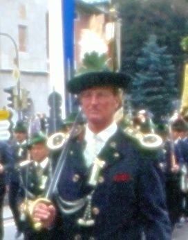 Ein Mann in einer traditionellen Militäruniform hält ein Schwert und marschiert in einer Parade mit anderen in ähnlichen Uniformen. Bäume und eine Ampel sind im Hintergrund.
