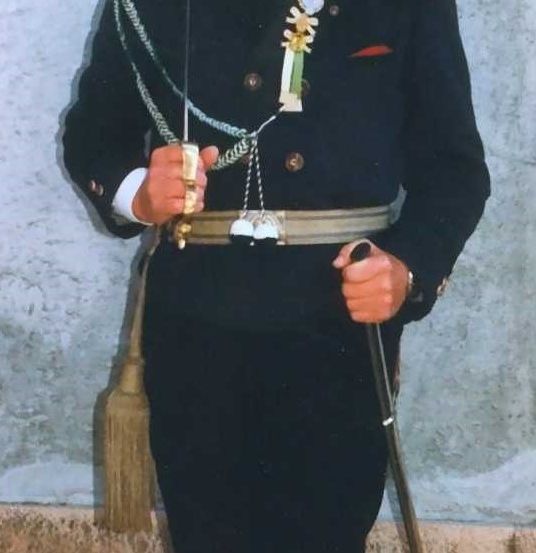 Eine Person in Uniform steht und hält ein Schwert und einen Schwertgürtel mit dekorativen Quasten. Sie trägt eine grüne Krawatte und verschiedene Medaillen auf der Brust.