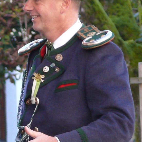 Ein Mann in einer Militäruniform mit grünen und roten Abzeichen, lächelnd, steht draußen.