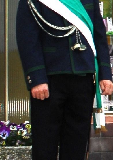 Ein Mann in einer traditionellen Uniform mit einer Schärpe steht vor einer Blumenvase.