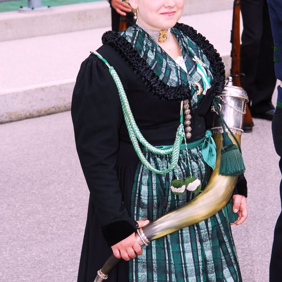 Eine Frau in einem grünen und schwarzen Kostüm mit einem Horn, lächelnd. Sie trägt eine Halskette, Ringe und eine Schärpe. Hinter ihr ist eine verschwommene Figur eines Mannes in schwarzen Hosen.