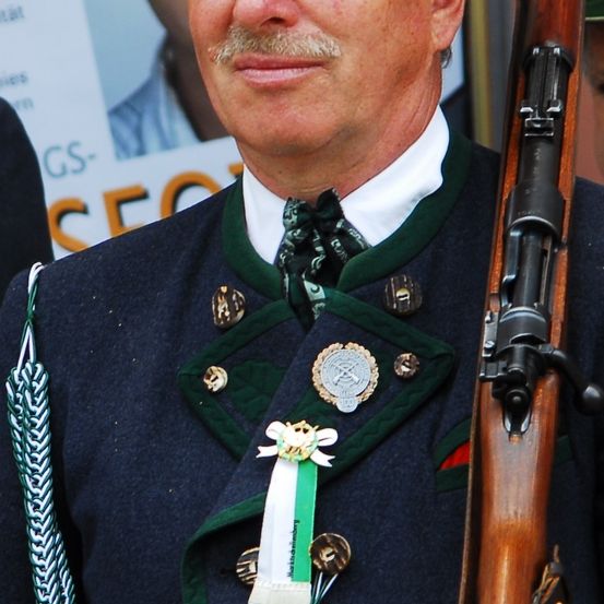 Ein Mann in einer grün-blauen Uniform mit einer Waffe lächelt. Er hat eine Abzeichen und ein grünes Band an seiner Jacke.