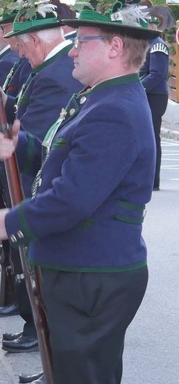 Eine Gruppe von Männern in traditionellen deutschen Uniformen steht in Formation und hält Gewehre. Sie tragen grüne Mützen mit Federn, blaue Jacken und schwarze Hosen. Sie befinden sich auf einer Straße mit Grünpflanzen im Hintergrund.