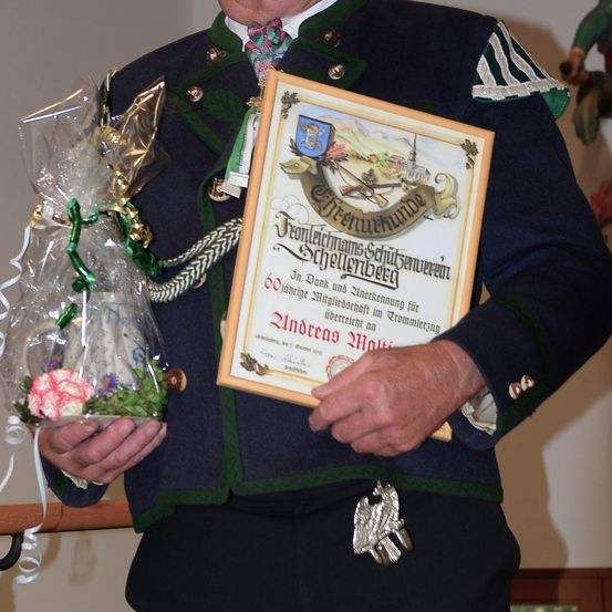 Ein Mann in traditioneller deutscher Kleidung hält ein Zertifikat und ein Geschenk in Plastikfolie. Das Zertifikat ist golden mit einer Schleife und Blumenarrangement.