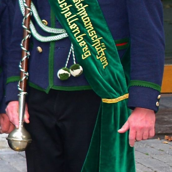Eine Person in einem blauen Uniform mit einem grünen Schal hält ein Metallinstrument. Der Schal hat goldenen Text, der 'Schellenbuerger' lautet.