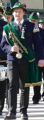 Ein Mann in einem grünen Schal und Hut läuft in einer Parade, hält einen Pokal und hat eine Marschkapelle hinter sich.