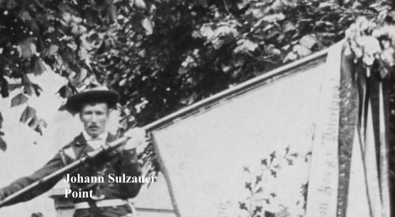 Ein Mann in Uniform steht auf einer Leiter und hält eine Fahne mit Text und Dekorationen. Er schaut nach vorne, und es gibt einen Baum im Hintergrund.