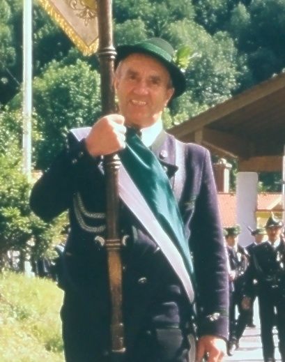 Ein älterer Mann in einem grünen Hut und einem grünen Schal hält einen Fahnenmast. Er ist von anderen uniformierten Personen umgeben, die auf einem Weg gehen.