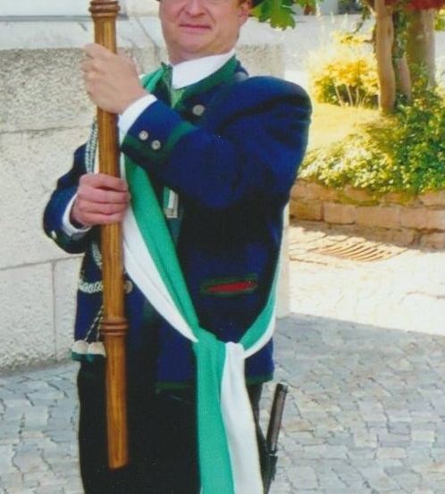 Ein Mann in traditioneller Kleidung steht auf einer Kopfsteinpflasterstraße und hält einen Holzstab. Er trägt einen grünen Hut, eine blau-grüne Jacke und eine weiße Schärpe.