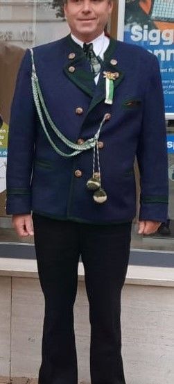 Ein Mann in einem blauen Jackett mit grünen Akzenten und einem grünen Hut mit Bändern steht draußen vor einem Gebäude.