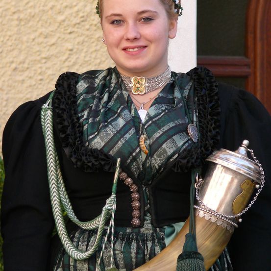 Eine lächelnde Frau in traditioneller Kleidung hält ein Horn, verziert mit einer silbernen Kette und Quaste. Ihr grün-schwarzes Kleid hat Rüschen und aufwendige Schmuckstücke. Sie steht vor einer hellen Wand.