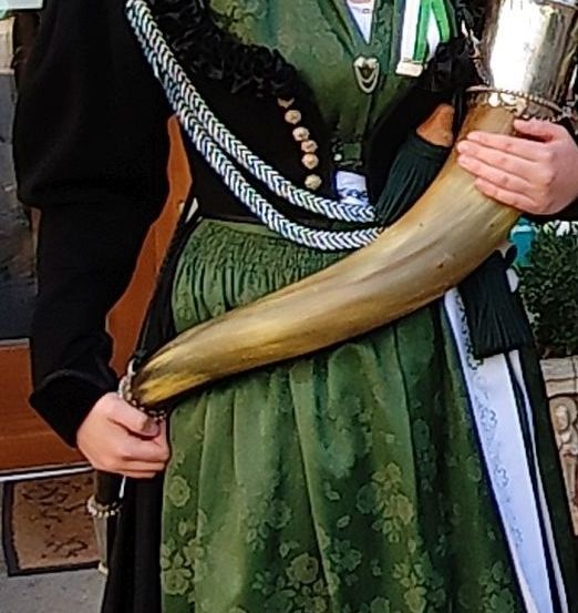 Eine Frau in einem grünen Kleid mit Blumenmustern, geschmückt mit Bändern, und hält ein großes Horn.