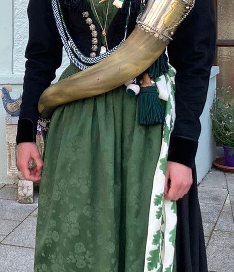 Eine Person in traditioneller Kleidung, die einen silbernen Horn mit Quaste hält, posiert vor einem Gebäude.