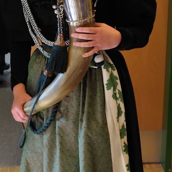 Eine Person in traditioneller Kleidung hält ein großes Horn mit aufwendigen Silberverzierungen. Das Horn hat eine Quaste und ist mit einem blauen Seil umwickelt.