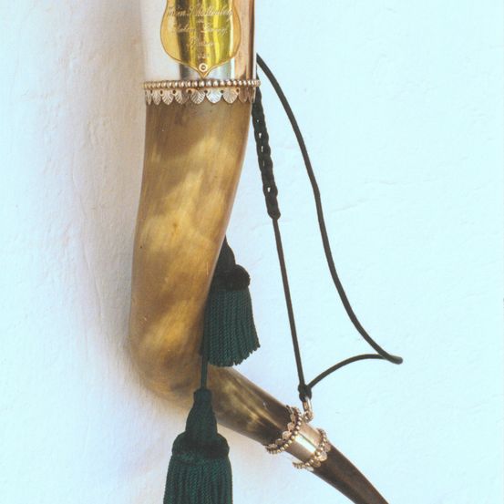 Ein goldener Horn mit silberner Einfassung ist an einer weißen Wand montiert. Es hat eine goldbraune Farbe und ist mit einem silbernen Band und einer grünen Quaste verziert.