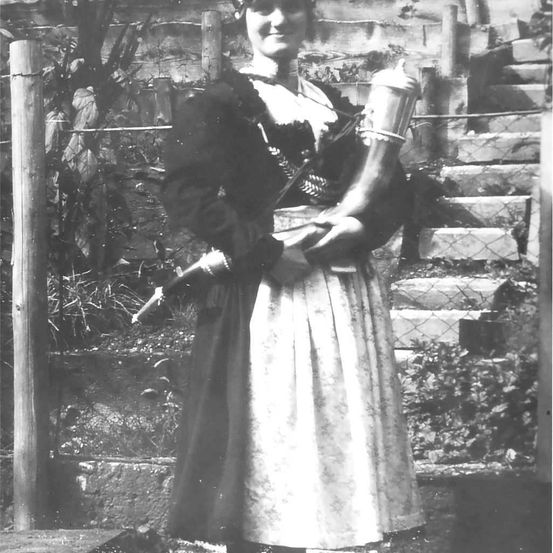 Ein Schwarz-Weiß-Bild einer Frau in traditioneller Kleidung, die eine Trompete in den Händen hält, steht in der Nähe eines Zauns und einer Treppe.