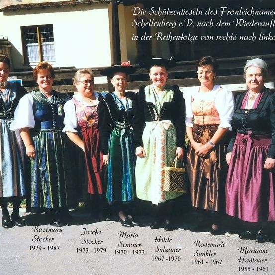 Sieben Frauen in traditioneller Kleidung stehen in einer Reihe, identifiziert durch ihre Namen und Jahre. Sie stehen vor einem Gebäude mit Treppen und einem Fenster. Der Text über ihnen lautet 'Die Schutzenlieseln des Frontleichnams Schellenberg e.V. nach dem Wiederauf.'