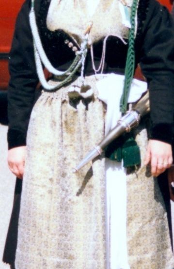 Eine Frau in traditioneller Kleidung mit einem Schwert und anderen Accessoires steht draußen. Hinter ihr ist ein rotes Fahrzeug zu sehen.