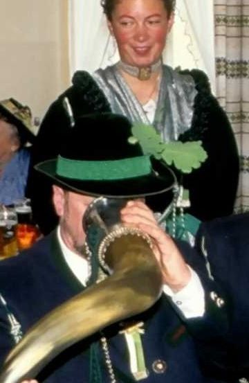 Ein Mann in traditioneller Kleidung mit einem Horn bläst es, während eine Frau in traditionellem Kleid hinter ihm lächelt.