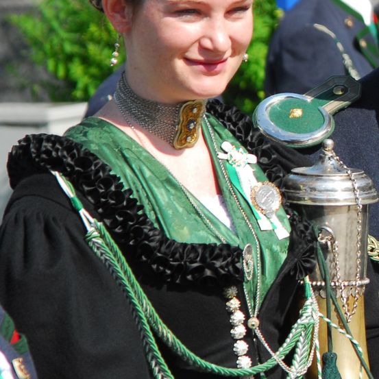Eine Frau in traditioneller grüner und schwarzer Kleidung mit dekorativen Ketten und Medaillen, lächelt mit einem silbernen Pokal in der Nähe.