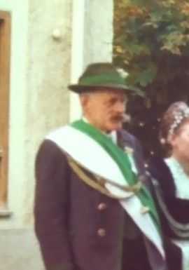 Ein älterer Mann mit grünem Hut und Schärpe geht neben einer Frau in traditioneller Kleidung vor einem Gebäude.