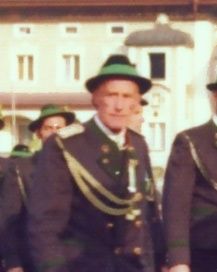 Ein älterer Mann in einer Militäruniform mit einem grünen Hut steht vor einem Gebäude. Er hat Medaillen auf der Brust.