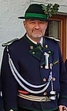 Ein Mann in traditioneller bayerischer Uniform steht vor einer weißen Wand. Er trägt einen grünen Hut mit einer Girlande, einen blauen Mantel mit Schulterklappen und eine grüne Schärpe.