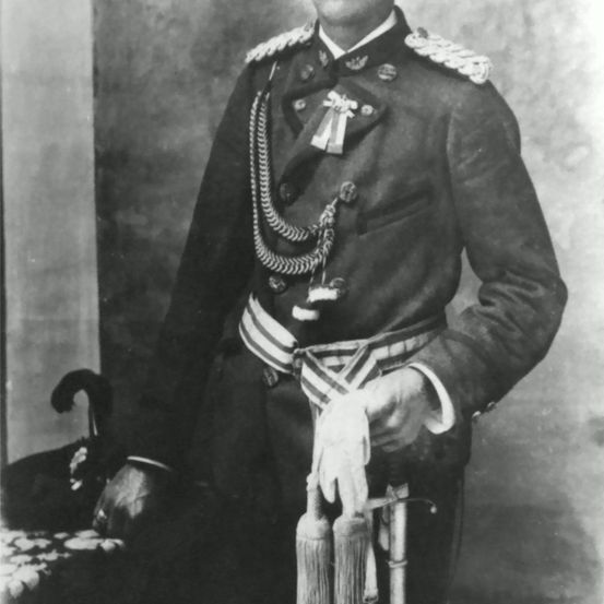 Ein Schwarz-Weiß-Foto eines Mannes in Militäruniform, der ein Schwert hält und vor einer Wand steht.