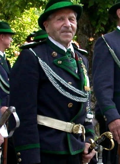 Ein älterer Mann in traditioneller Uniform steht vor einem Baum und hält ein Schwert, mit mehreren anderen in Uniform hinter ihm.