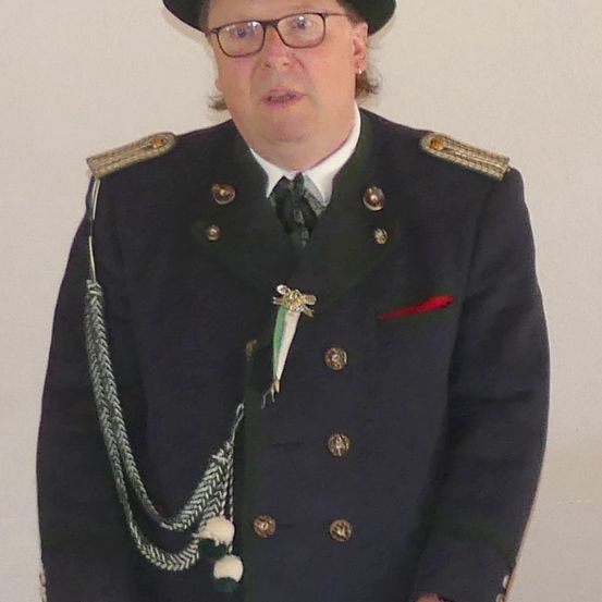 Ein Mann in einer dunkelblauen Uniform mit goldenen Knöpfen, einer schwarzen Krawatte und einem Hut steht an einer weißen Wand. Er hat eine Medaille auf der Brust.