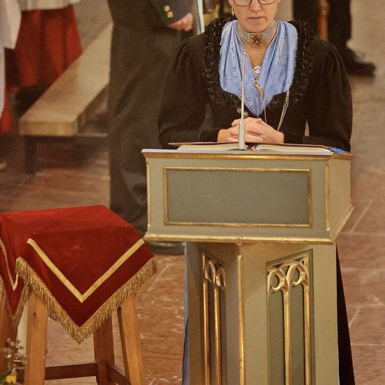 Eine Frau steht an einem Rednerpult und liest aus einem Buch, während andere in der Nähe stehen. Hinter ihr ist eine Bank und eine Person in schwarzen Schuhen zu sehen.