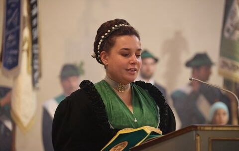 Eine Frau steht an einem Podium mit einem Mikrofon und hält ein Buch. Sie trägt ein grünes Kleid und einen schwarzen Umhang. Hinter ihr sind verschwommene Figuren zu sehen, darunter zwei Männer in Hüten und eine Frau mit einer weißen Kappe.