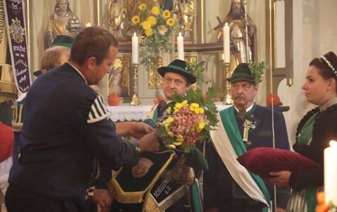 Eine Gruppe von Menschen in traditioneller Kleidung versammelt sich in einer Kirche. Zwei Männer halten einen Blumenstrauß, während ein dritter Mann etwas in eine Vase legt.