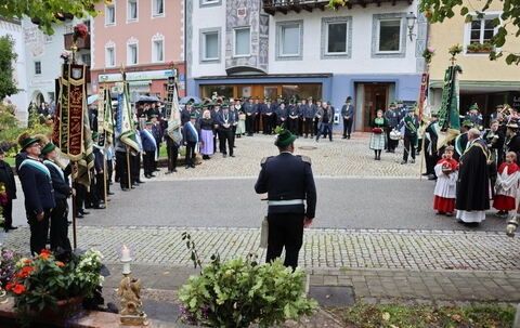 Eine Gruppe von Menschen in Uniform und traditioneller Kleidung steht auf einer Kopfsteinpflasterstraße mit Fahnen, Blumen und Gebäuden im Hintergrund.