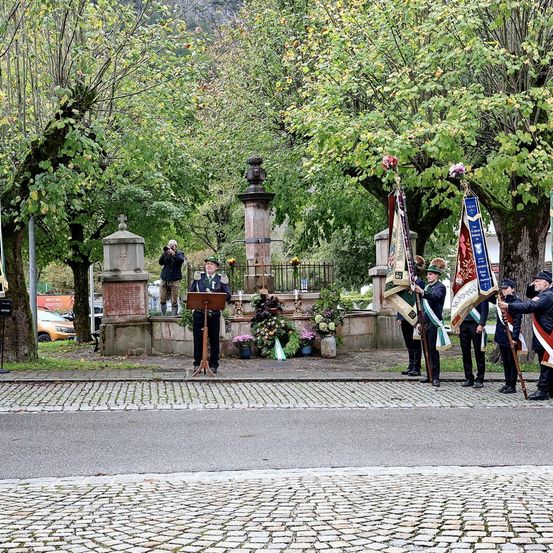 Eine Gruppe von Menschen in Uniform, die Fahnen halten, steht in einem Park, ein Mann ist auf einem Podium. Dahinter befinden sich Blumen, Bäume und ein Springbrunnen. Ein Mann macht ein Foto.