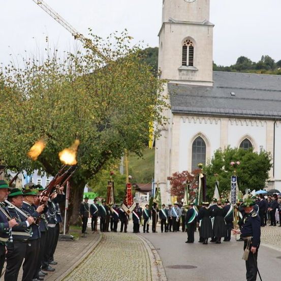 Eine Gruppe von Männern in Uniform und Hüten steht vor einer Kirche und feuert Gewehre ab. Im Hintergrund befinden sich Bäume, Fahnen und ein Kran.