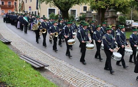 Eine Musikkapelle in Uniformen tritt im Freien auf. Sie tragen verschiedene Instrumente, darunter Trompeten, Posaunen und Trommeln. Die Kapelle läuft auf einer gepflasterten Straße.