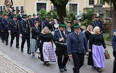 Ein Umzug mit Menschen in traditioneller deutscher Kleidung, einschließlich Hüten, die eine Kopfsteinpflasterstraße hinuntermarschieren. Einige tragen Gewehre, während andere Handtaschen haben.