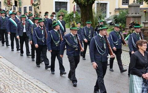 Eine Gruppe von Männern in Uniform mit grünen Hüten und Gewehren marschiert die Straße entlang. Eine Frau geht an der Seite.