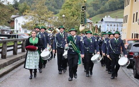 Eine Musikkapelle in traditionellen Uniformen tritt auf einer Straße auf. Eine Frau vorn trägt ein rotes Kissen, während andere Trommeln und Trompeten spielen. Im Hintergrund befinden sich Gebäude und Bäume.