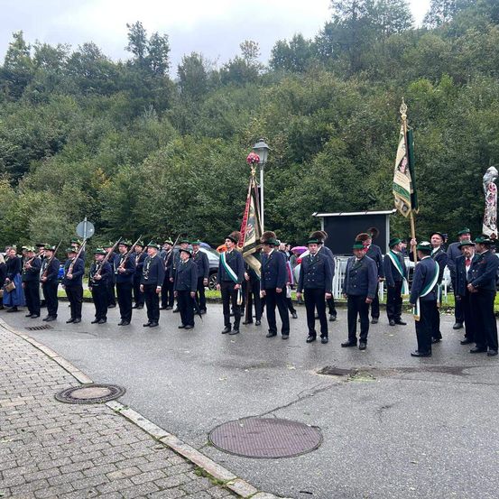 Eine Gruppe uniformierter Personen in Hüten und mit Gewehr bewaffnet steht auf einer Straße, mit einem Auto und Grün im Hintergrund.
