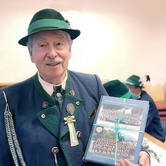 Ein älterer Mann in traditioneller grüner Hut und blauem Mantel hält ein eingewickeltes Buch und lächelt. Im Hintergrund sitzen andere Personen in ähnlicher Kleidung auf Stühlen.