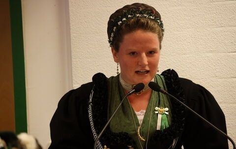 Eine Frau in traditioneller Kleidung spricht in ein Mikrofon bei einer Veranstaltung. Sie trägt eine Blumenkrone und eine grüne Schleife.